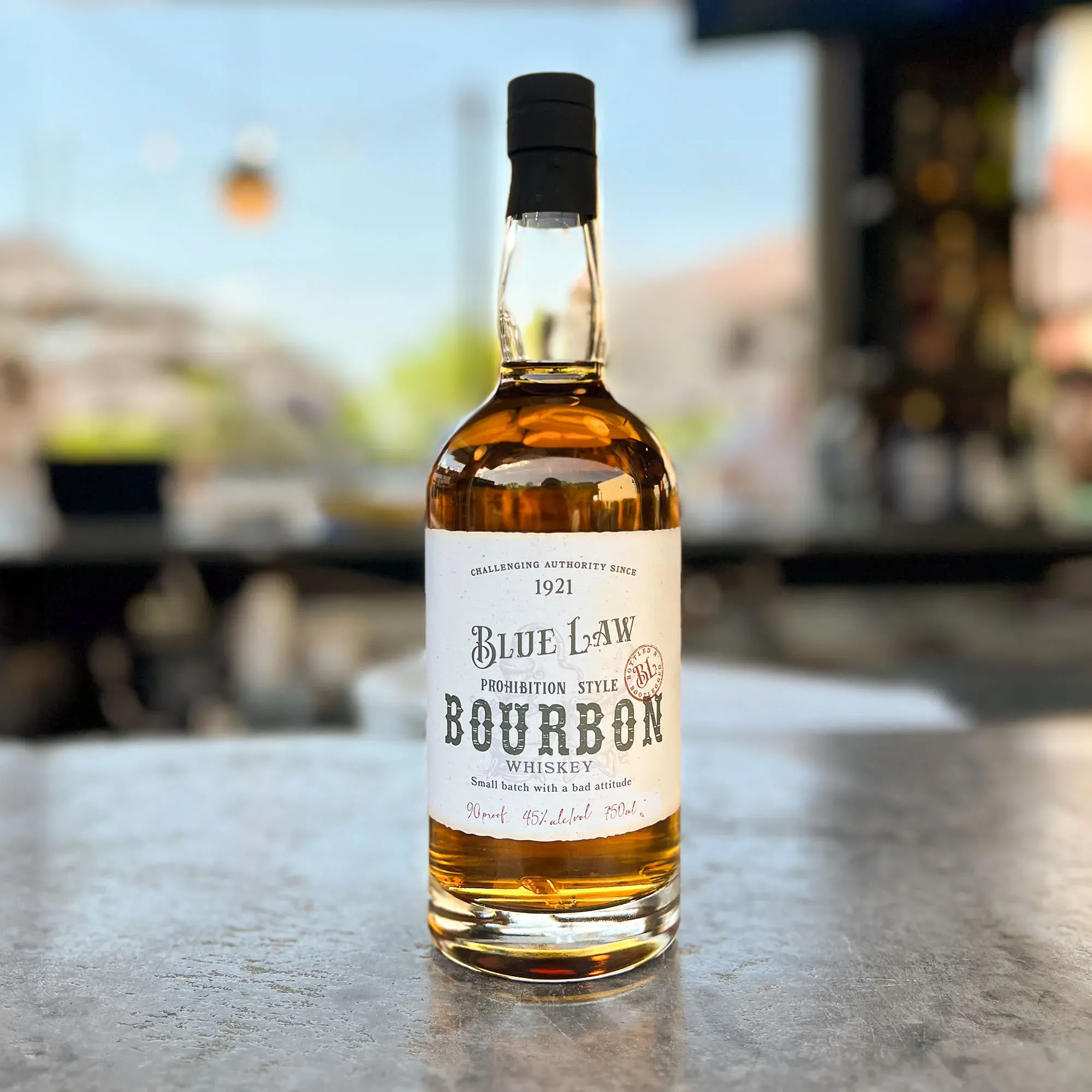 Blue Law Bourbon bottle in sharp focus on a patio table with three friends drinking cocktails.