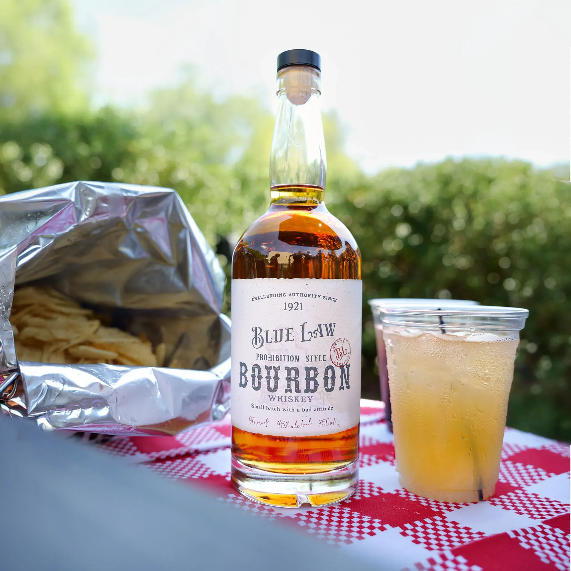 Blue Law Prohibition Style Bourbon bottle on a red checkered tablecloth next to a bag of chips.