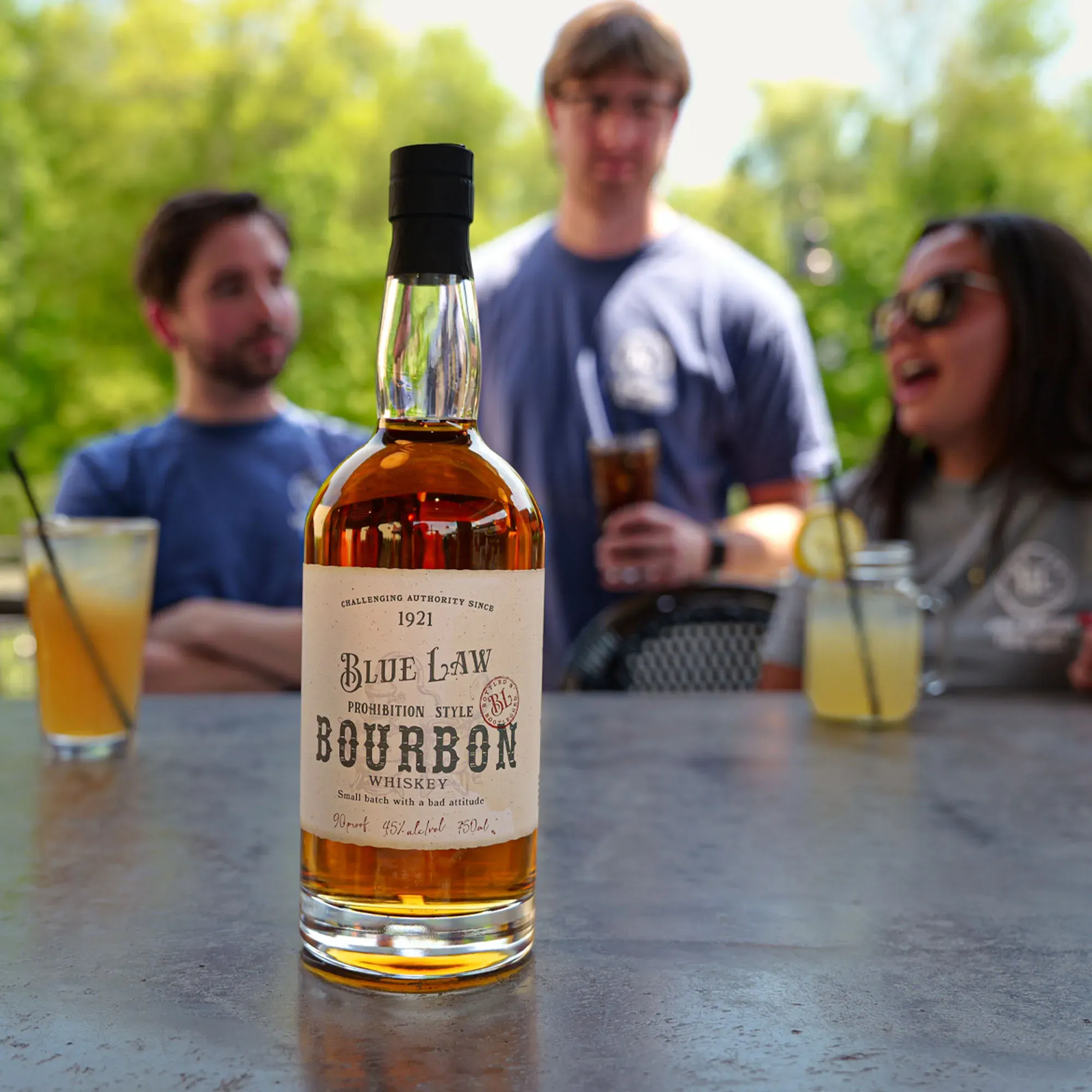 Blue Law Prohibition Style Bourbon Whiskey 750ml bottle on a metal bar counter with a outdoor bar in the background.