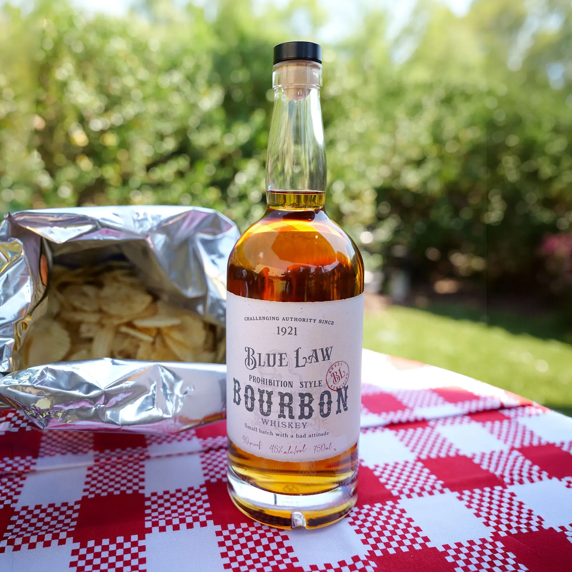 Blue Law Bourbon bottle on a red gingham picnic tablecloth beside an open bag of potato chips and an iced highball in a plastic cup.
